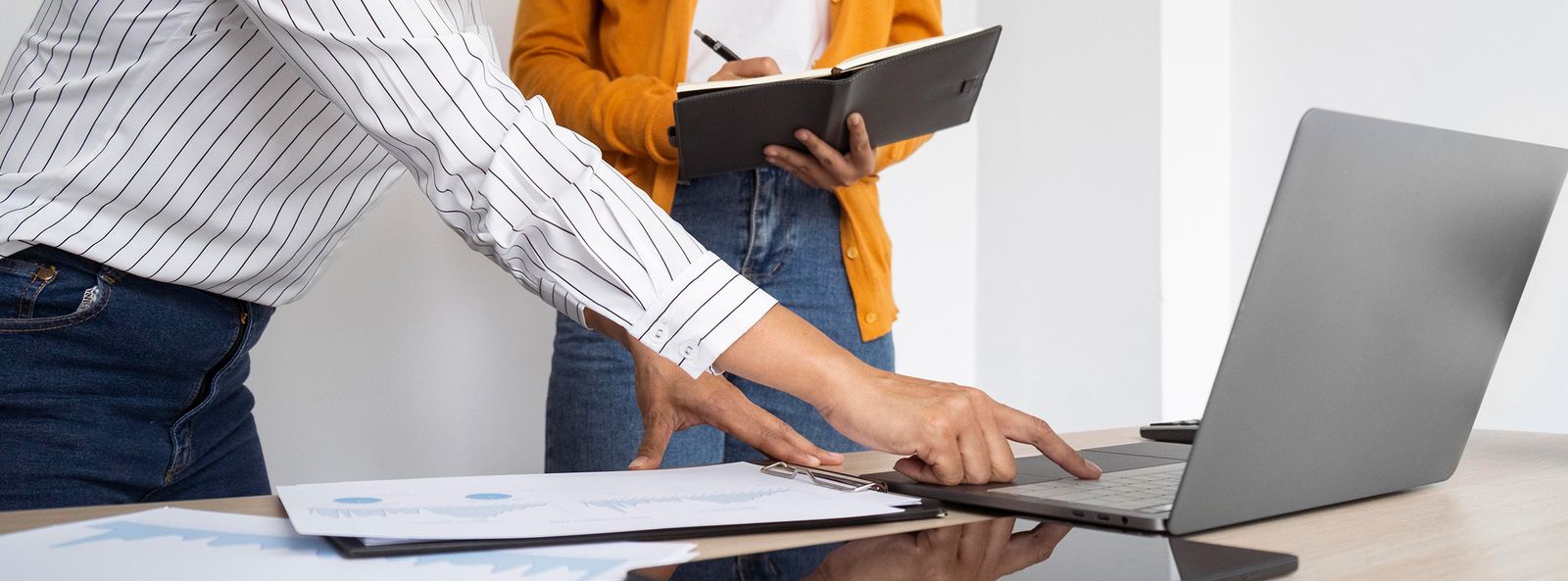 Una persona señala la pantalla de una notebook mientras otra toma notas de pie, con gráficos impresos sobre la mesa