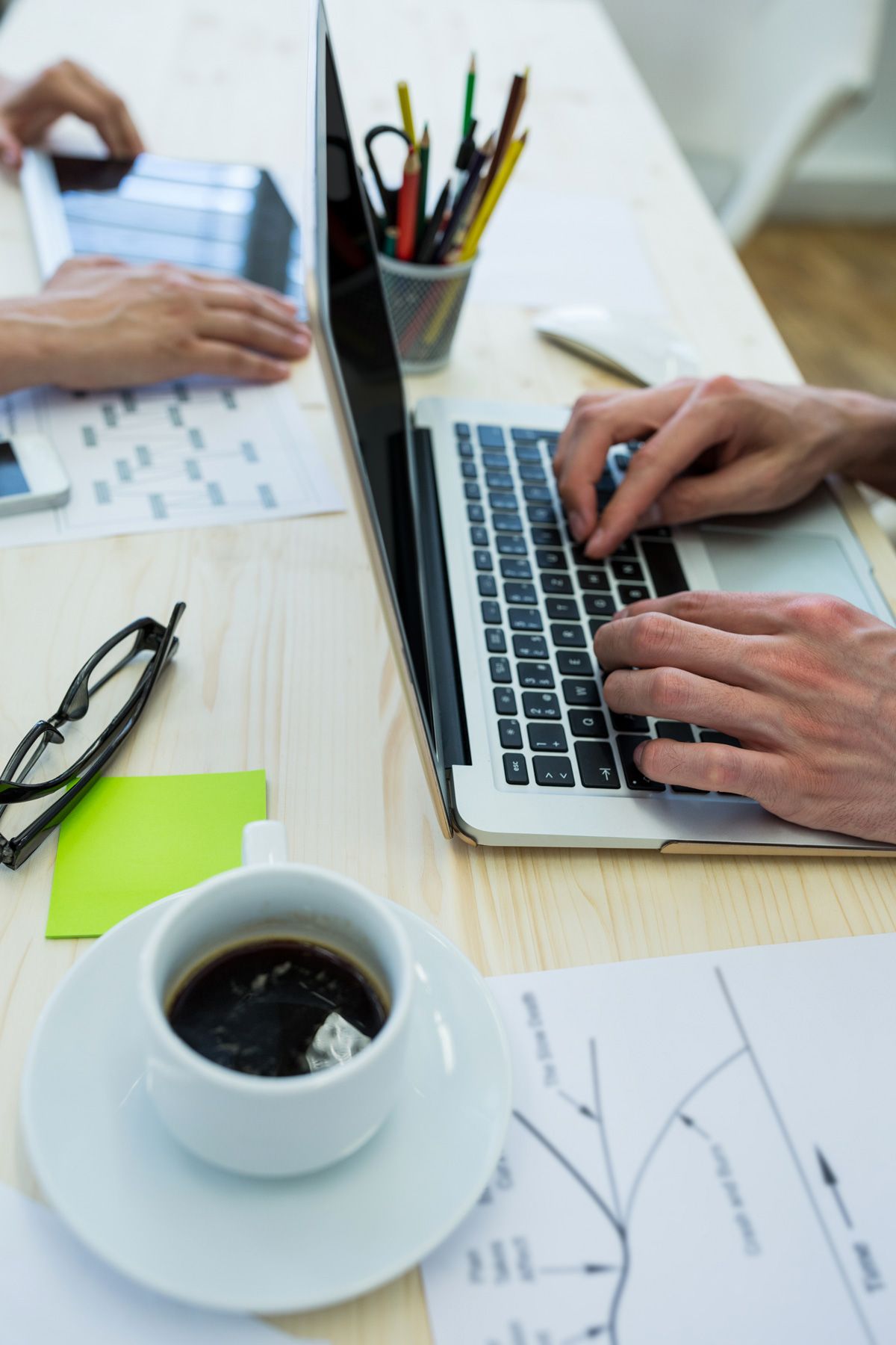 Manos escribiendo en una notebook sobre una mesa clara, con una taza de café, un portalápices y papeles con esquemas