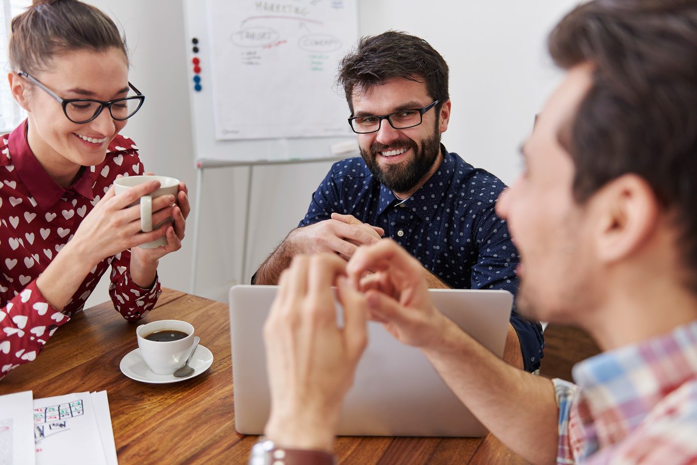 Tres personas sonriendo en una mesa de trabajo con una notebook y café, frente a un pizarrón